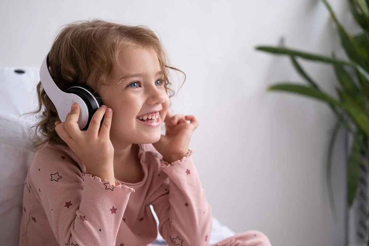Smiling child wearing headphones and a pink pajama with stars, sitting indoors near a plant.