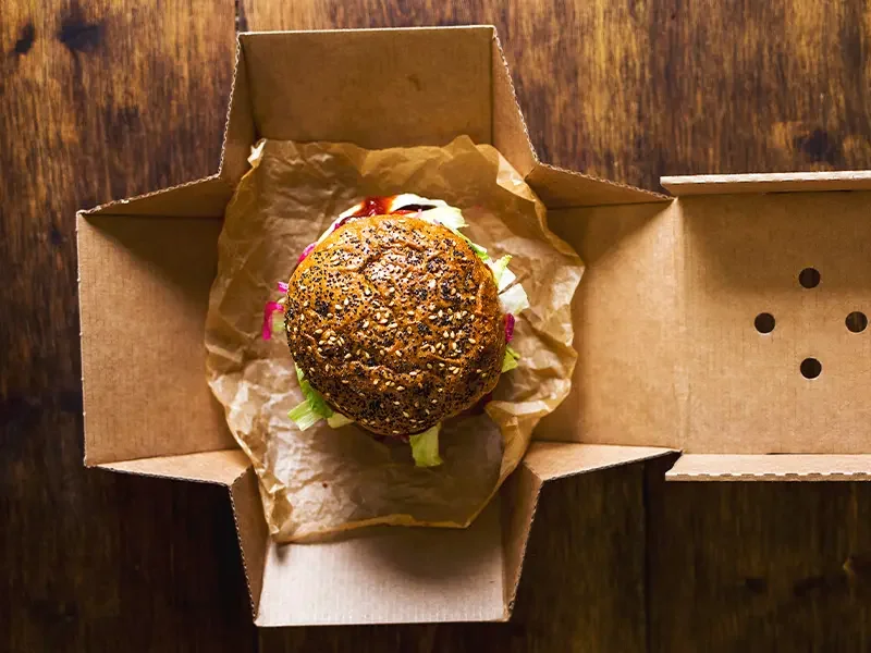 Overhead view of a gourmet burger in eco-friendly cardboard packaging, representing how menu development services design, test, and deliver market-ready food concepts that balance taste, sustainability, and consumer appeal.