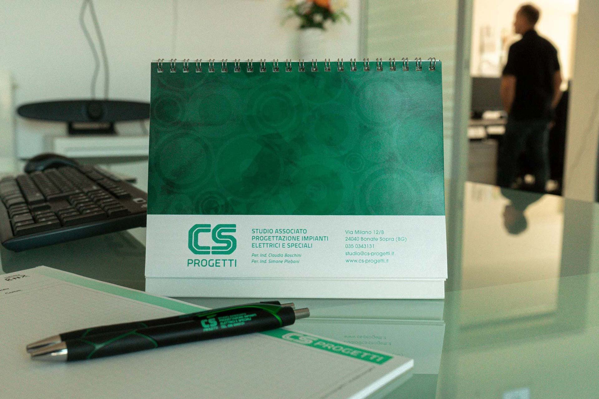 Un calendario da tavolo e una penna posati su una scrivania con altri oggetti, sullo sfondo ci sono una tastiera per computer e una persona che lavora in un ufficio.