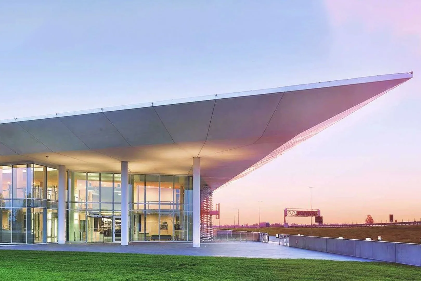 Edificio moderno con ampio tetto sospeso e grandi finestre, situato in un'area aperta con prato verde e un cielo al tramonto.