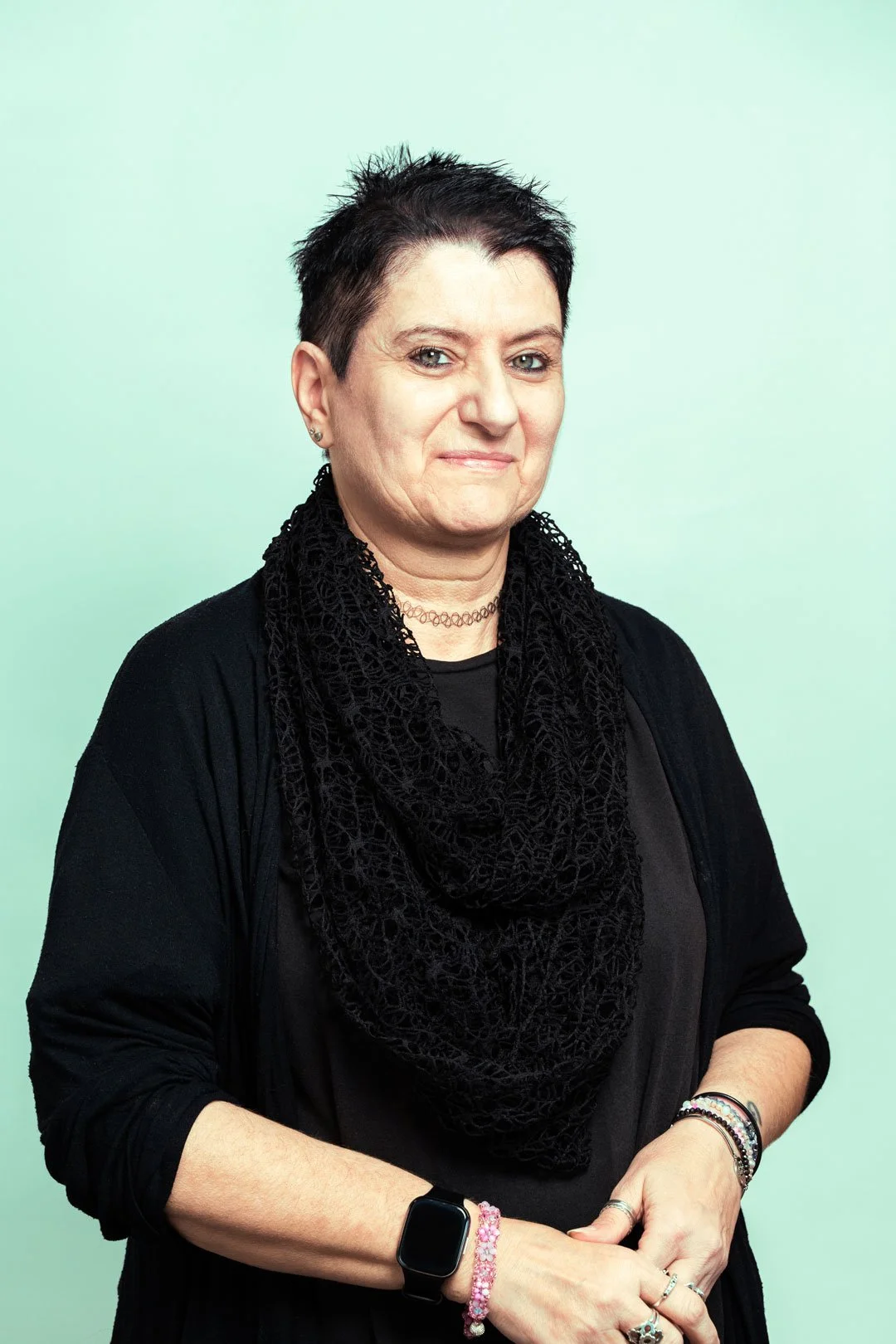 Donna con capelli corti e vestito nero, con diversi braccialetti e un orologio intelligente, posa davanti a uno sfondo verde chiaro.