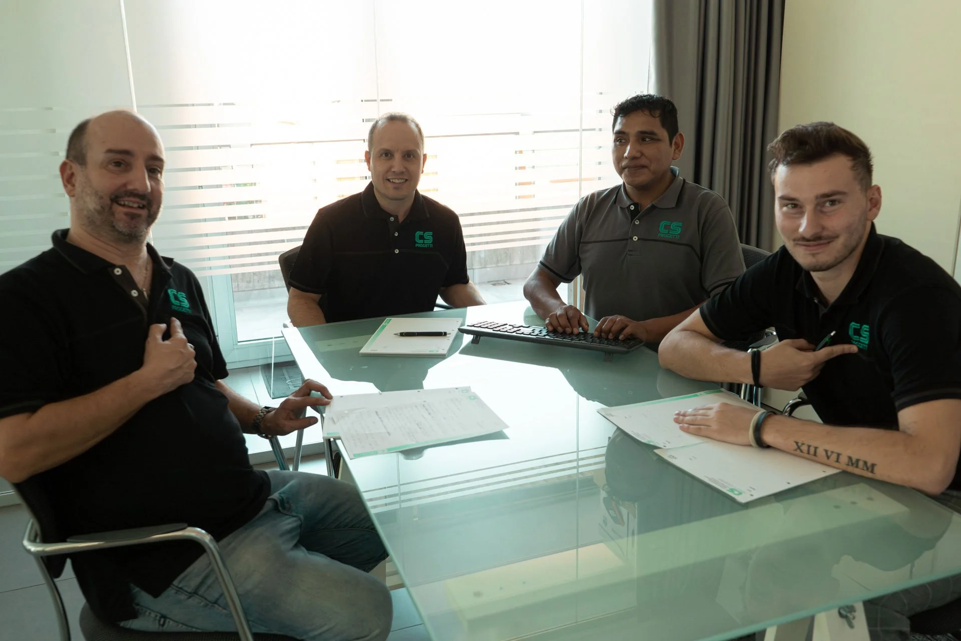 Quattro uomini in riunione seduti attorno a un tavolo di vetro, indossando magliette nere o grigie con il logo 'CS PROGETTI', con documenti e una tastiera.