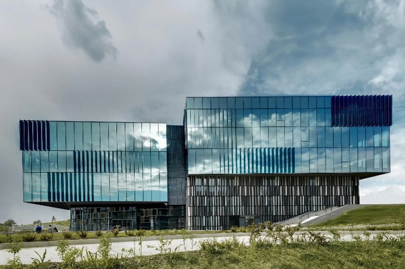 Edificio moderno con facciata in vetro riflettente, situato su un prato verde con percorso pedonale, sotto un cielo nuvoloso.