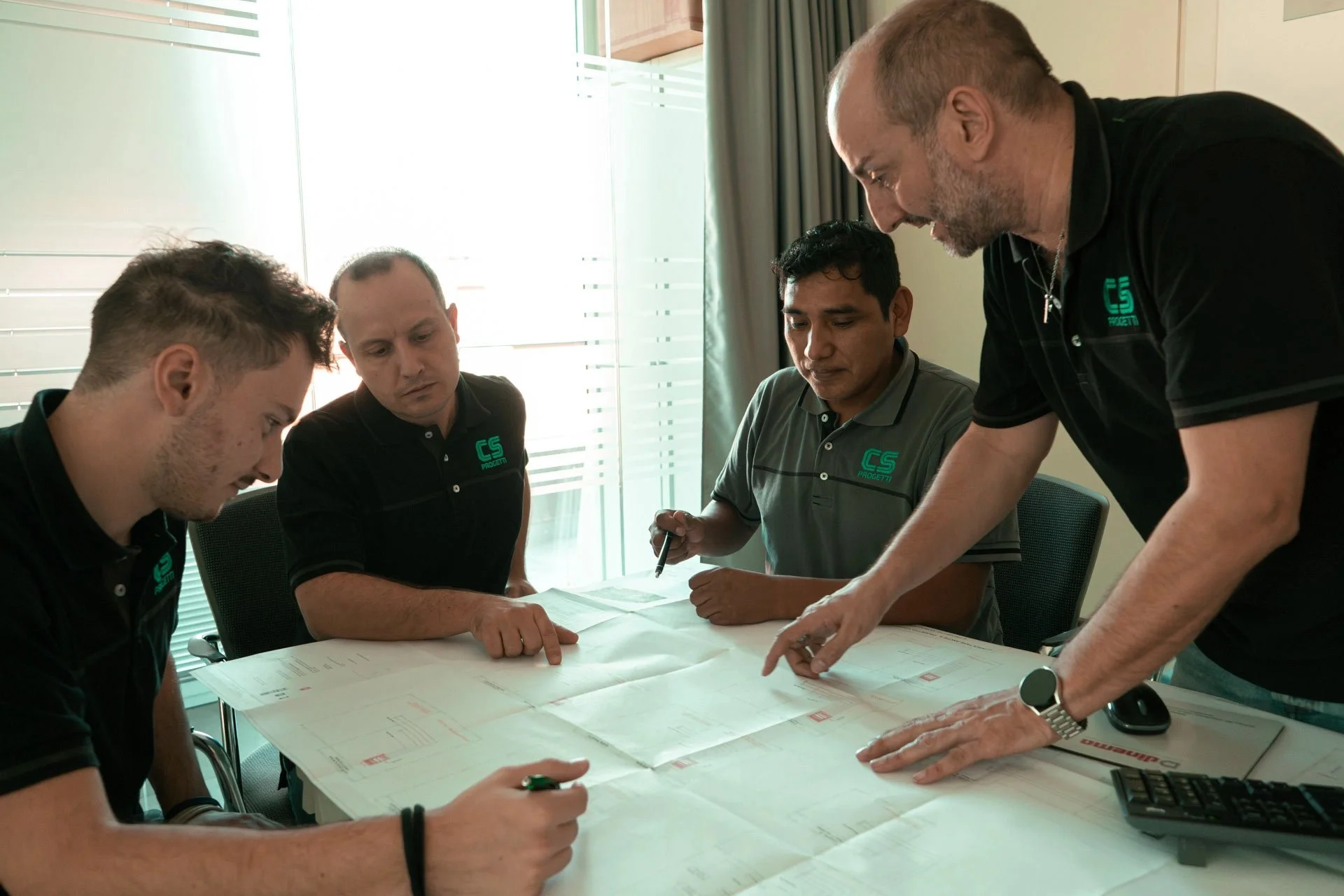 Quattro uomini seduti e in piedi intorno a un tavolo con dei progetti, che discutono e prendono appunti durante una riunione in ufficio.