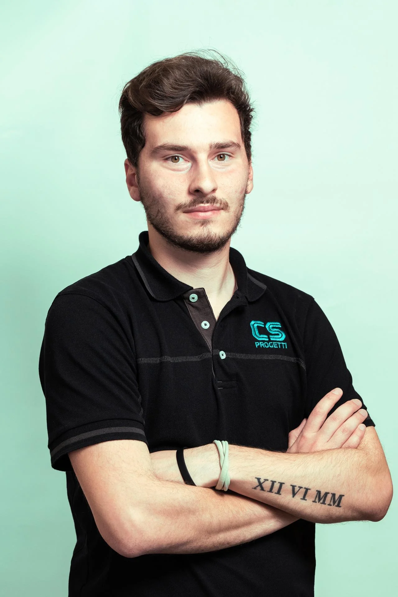Ragazzo con capelli castani, barba leggera, che indossa una polo nera con logo blu sulla sinistra, con braccia incrociate, sullo sfondo verde chiaro.