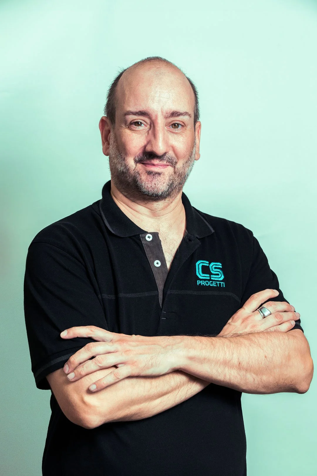 Uomo con barba, in piedi con le braccia incrociate, indossa una maglietta nera con il logo 'CS Progetti' di colore blu, sfondo verde chiaro.