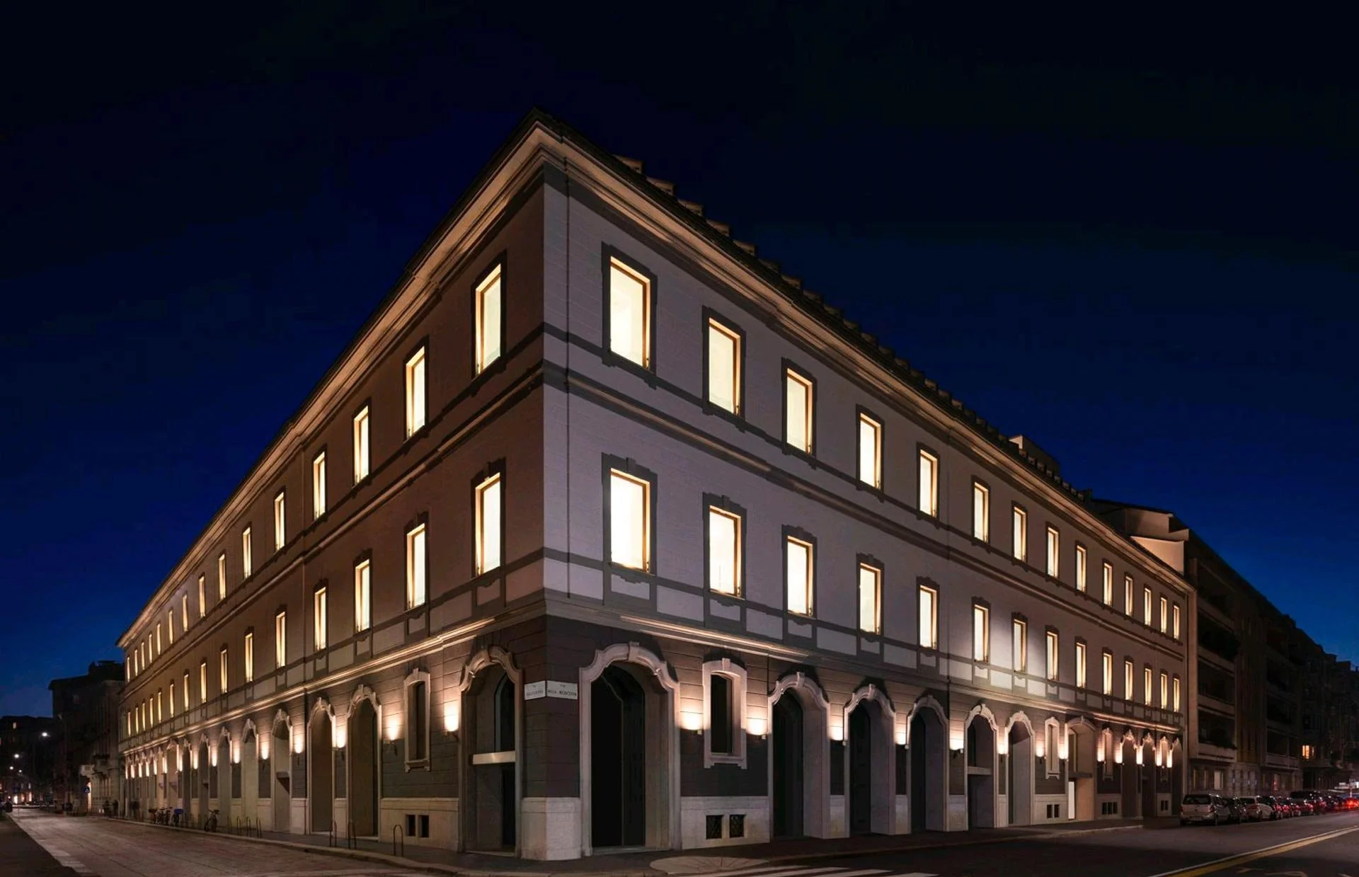 Edificio storico di cinque piani con illuminazione esterna, luci accese nelle finestre, situato in una strada urbana di sera.