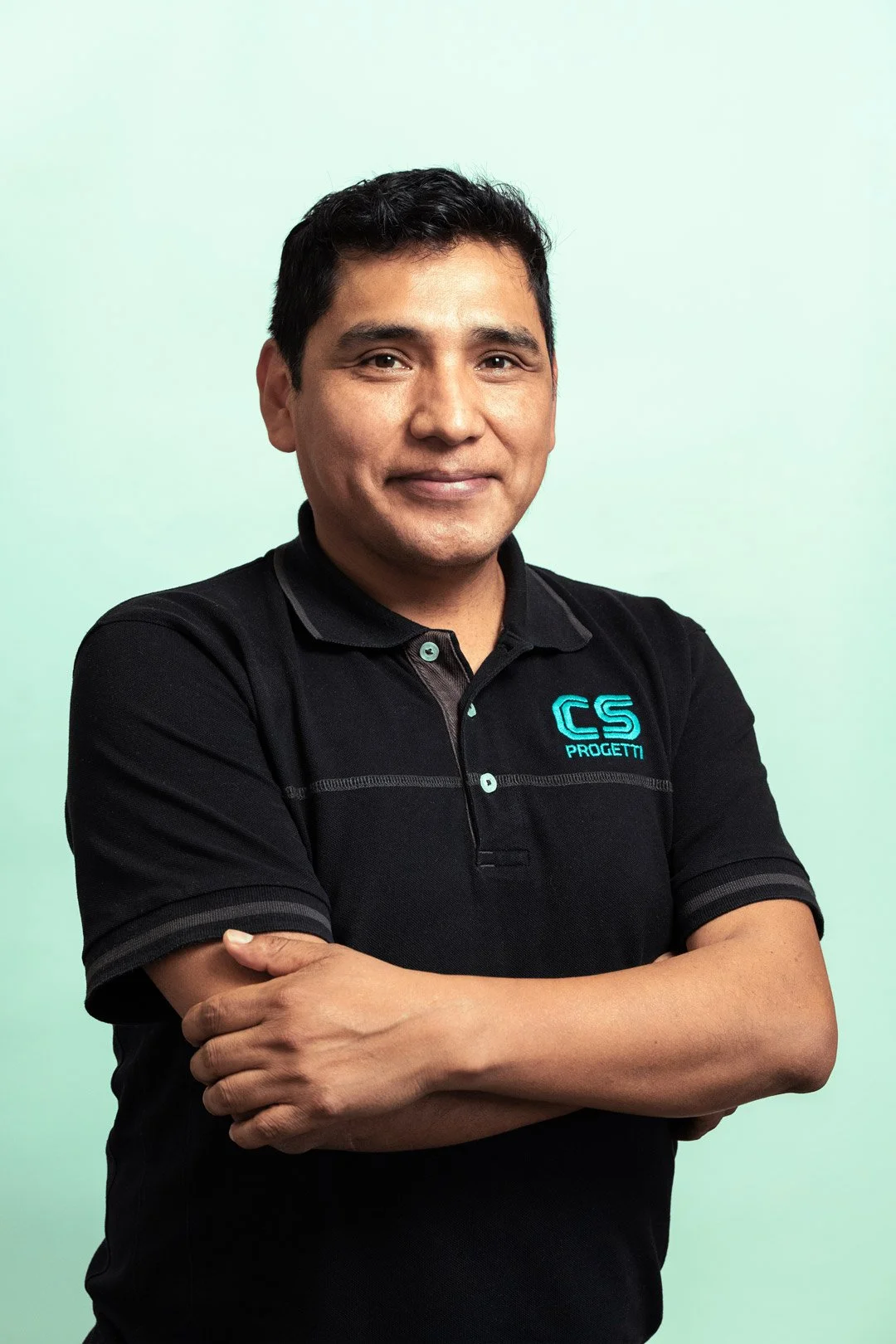 Uomo sorridente con capelli scuri, indossa una polo nera con logo 'CS Progetti' blu, posa con le braccia incrociate davanti a uno sfondo verde chiaro.