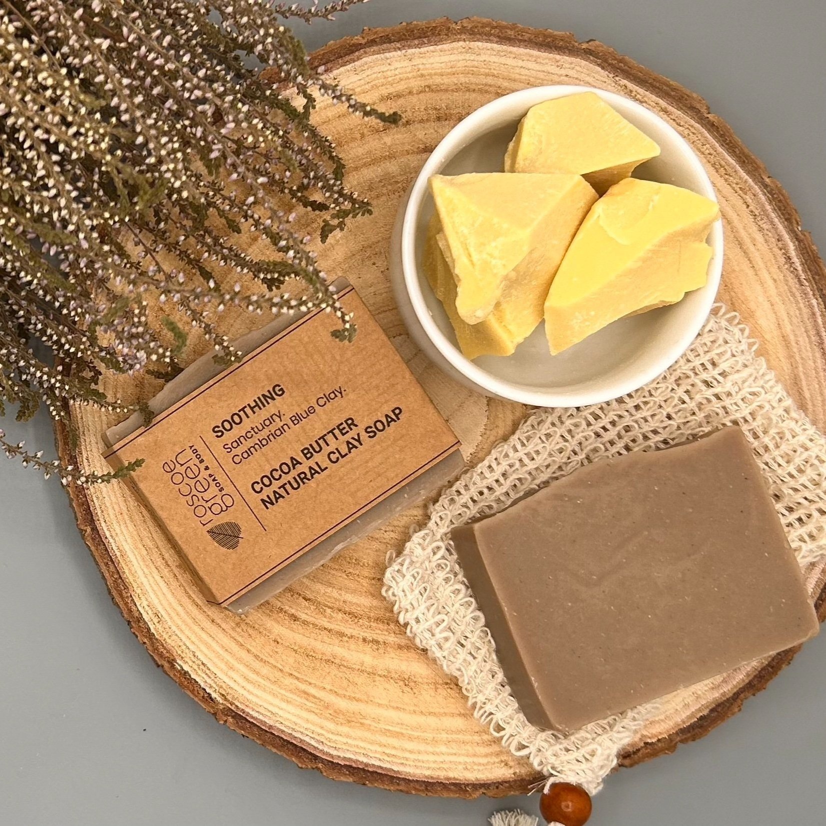 Sanctuary and Cambrian Blue Clay - Cocoa Butter and Clay Natural Soap bar placed on a wood slice with cocoa butter and heather.