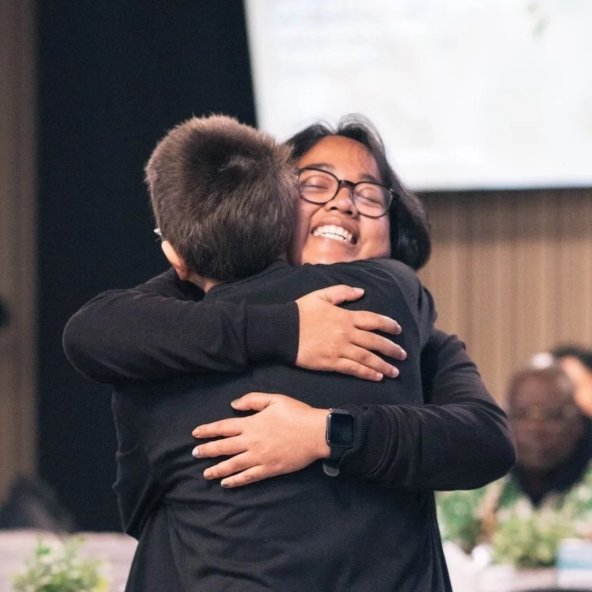 Two people hugging, smiling happily, in an indoor setting with other people in the background.