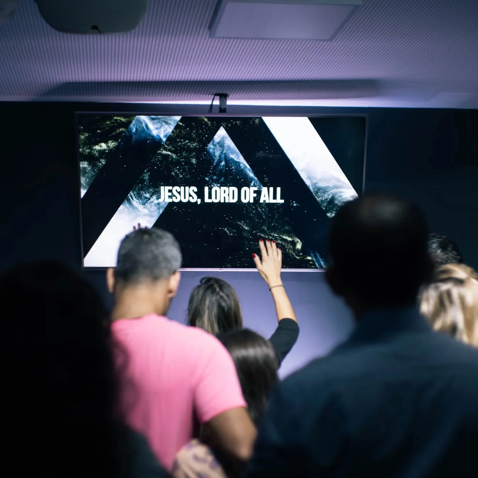 A group of people in a dark room watching a presentation on a large screen that displays the words 'JESUS, LORD OF ALL' over a mountain scene.