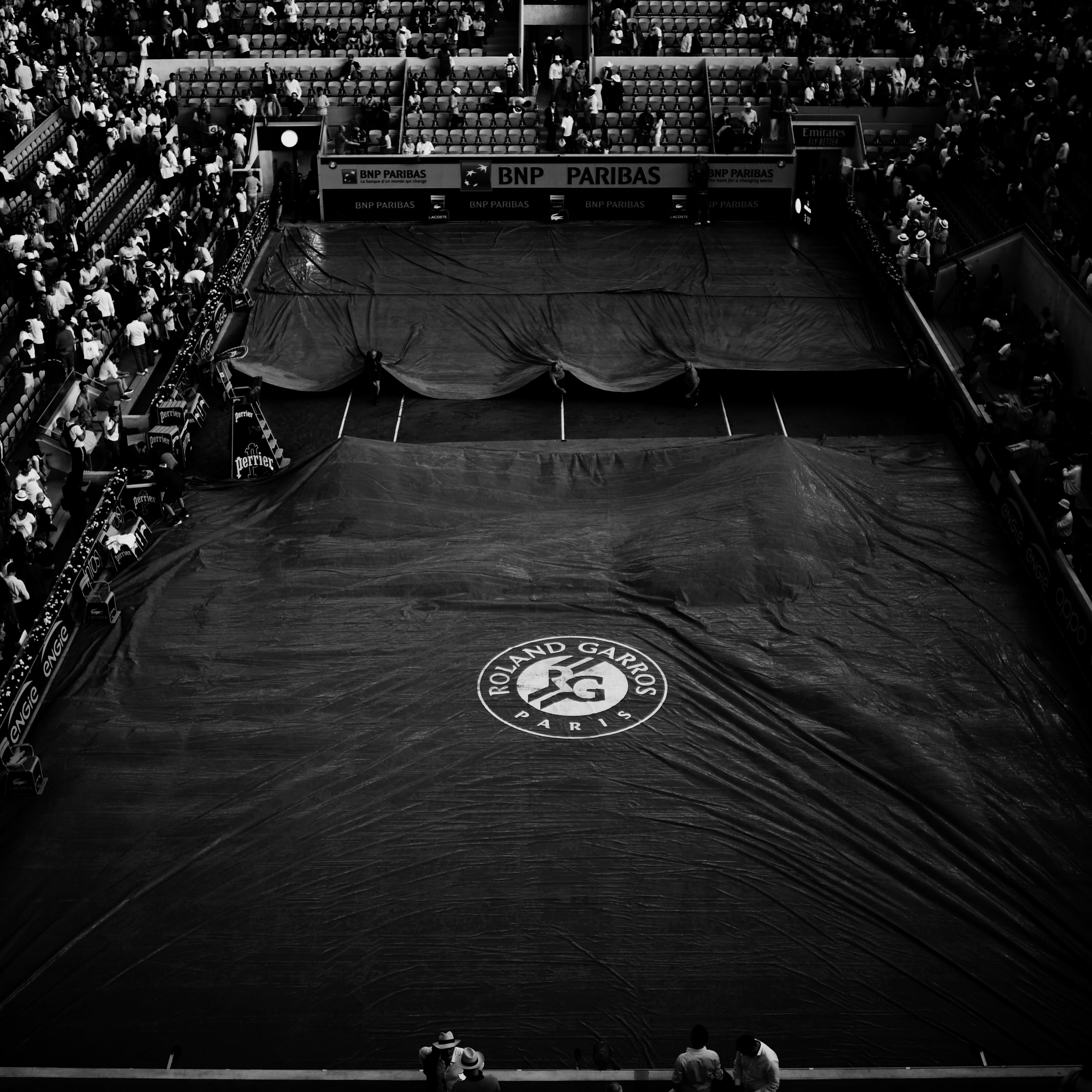 An indoor tennis court with a dark cover and a Roland Garros logo, surrounded by spectators, with some people rolling out the cover. court vibes only