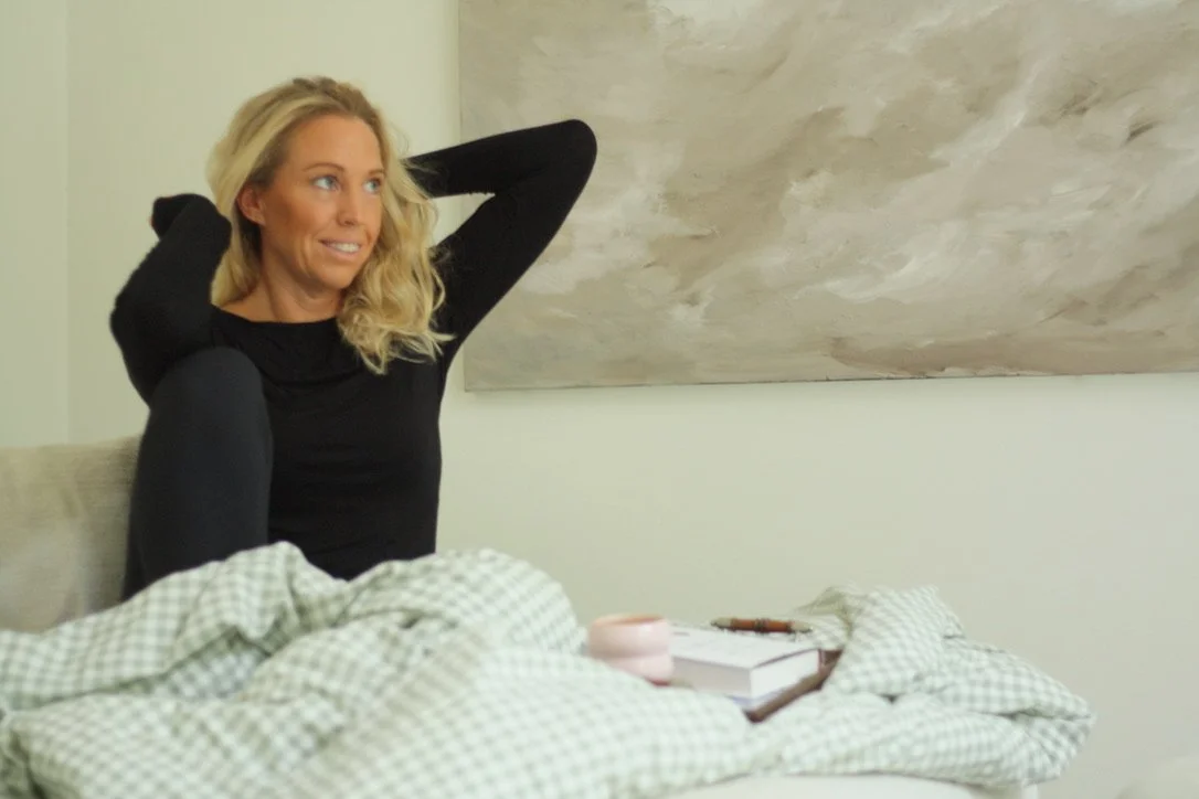 En kvinna sitter på en säng och tänker, med händerna bakom huvudet, i ett sovrum med ett stort konstverk på väggen och ett bord med några böcker och en ljus rosa ljuslykta.