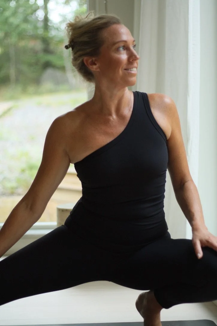 En kvinna i svart träningskläder som sitter i en yogaställning vid ett fönster med grönt utomhus.