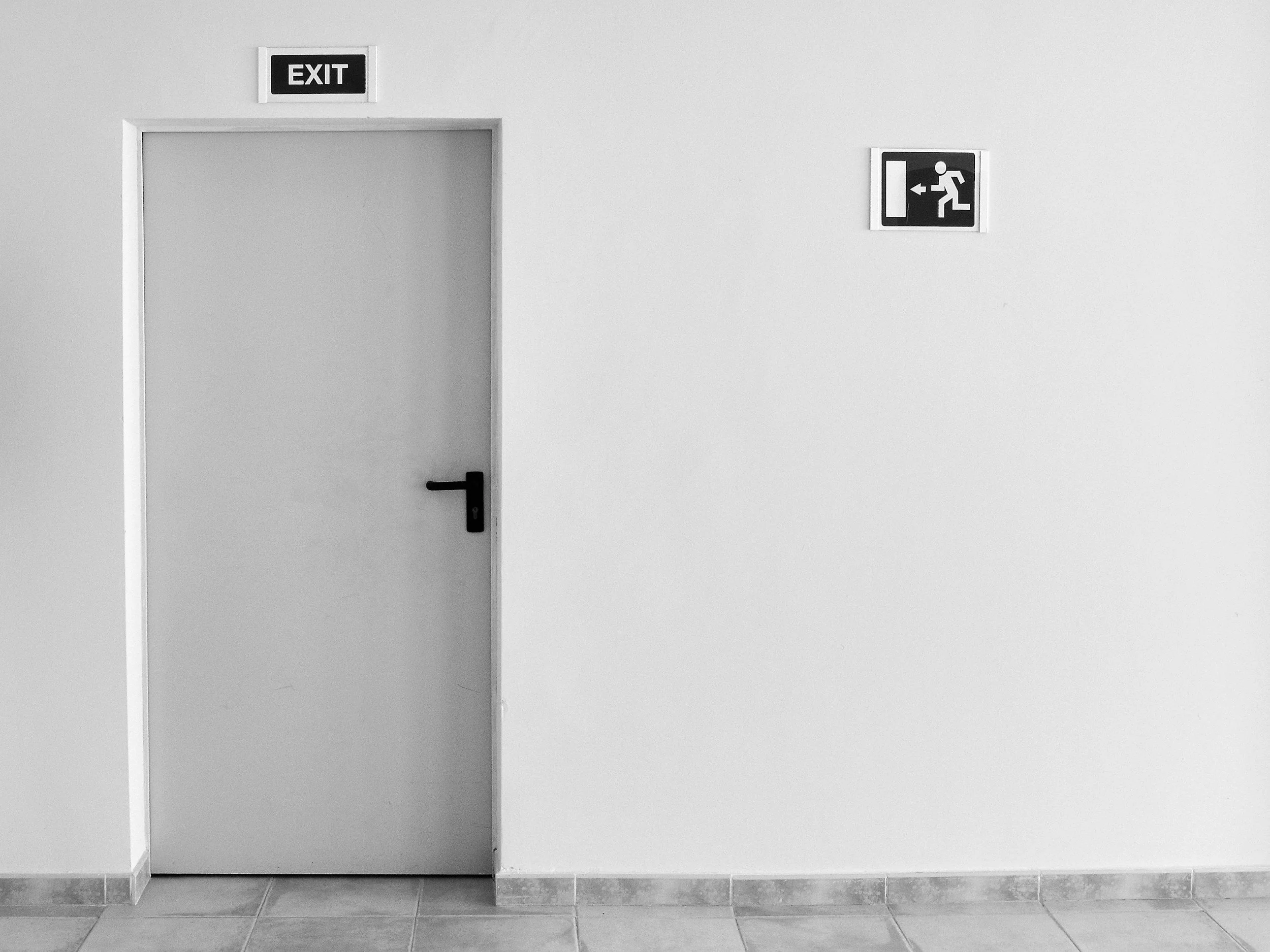 White door with "EXIT" sign above, black handle, and emergency exit symbol on the wall.