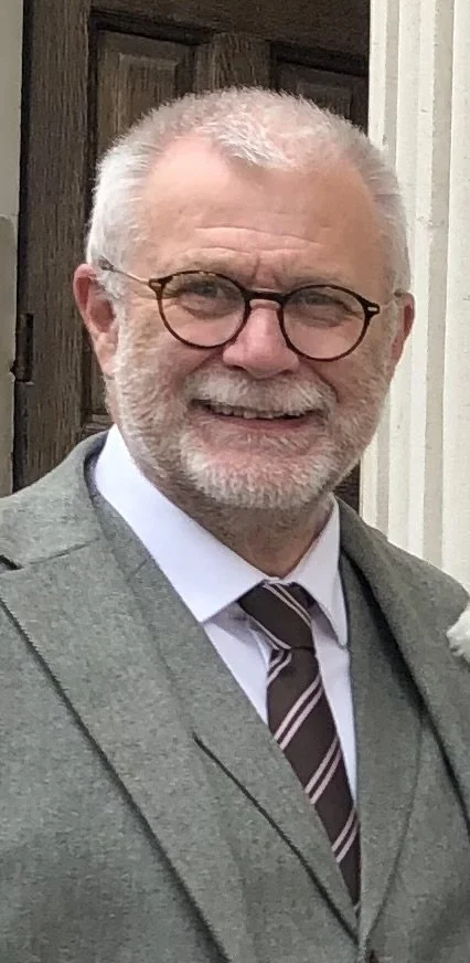 Elderly man wearing gray suit, tie, and glasses, smiling outdoors, standing in front of a door.