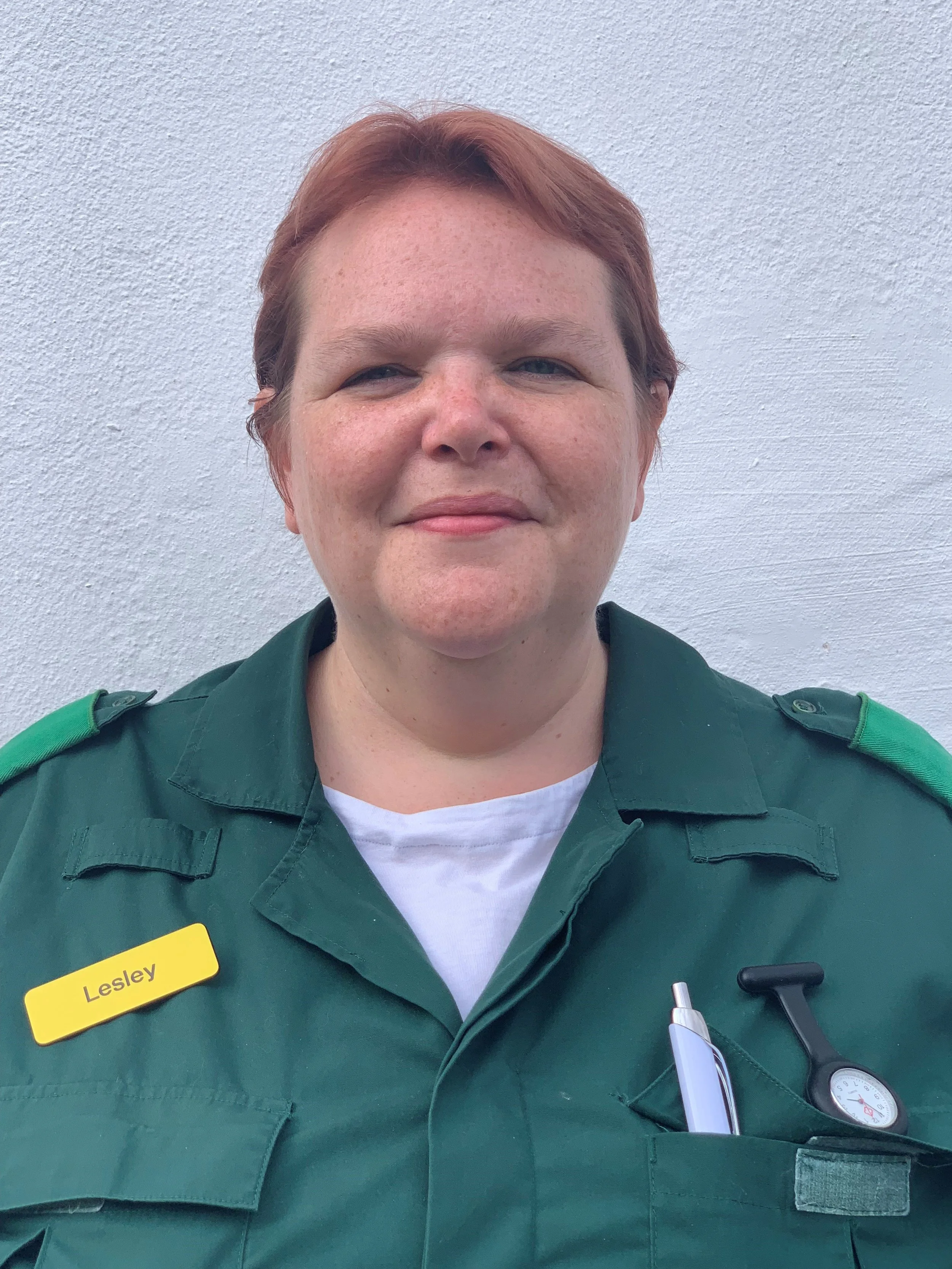 Person in a green uniform with a yellow name badge reading "Lesley," standing against a white background; features a pen and a watch pin on the uniform.
