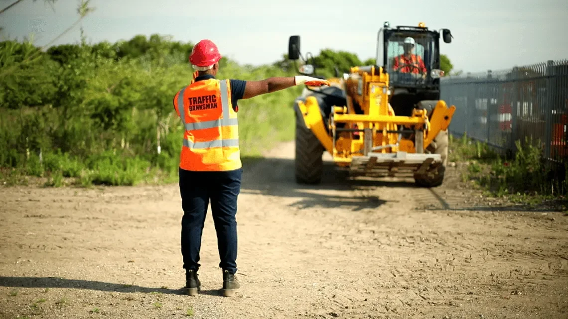 Traffic control worker in safety vest and red helmet directing construction equipment with a green shift in a construction zone.