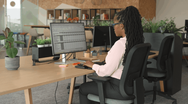 A woman working at a computer in an office with plants and bookshelves in the background.