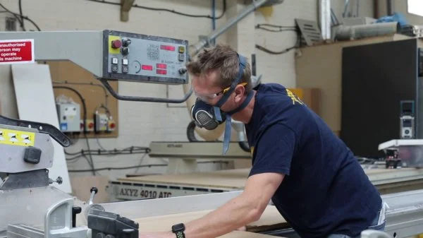 A man wearing safety goggles and a face mask working with a large industrial machine in a workshop.