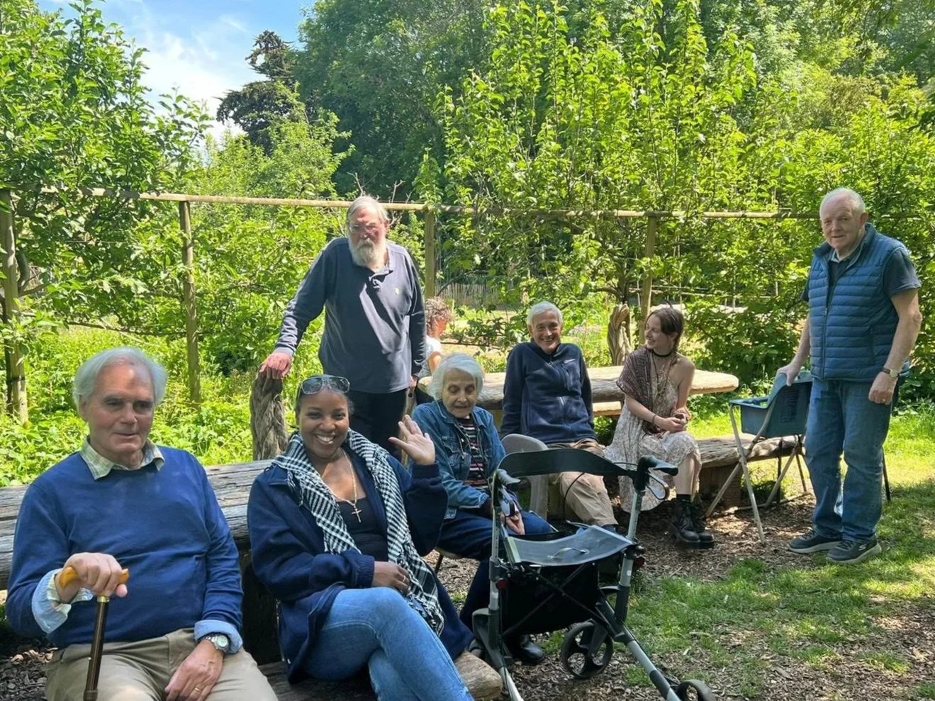 A group of seven elderly and middle-aged people, including one with a walker, sitting on benches outdoors in a lush garden, smiling and socializing on a sunny day.