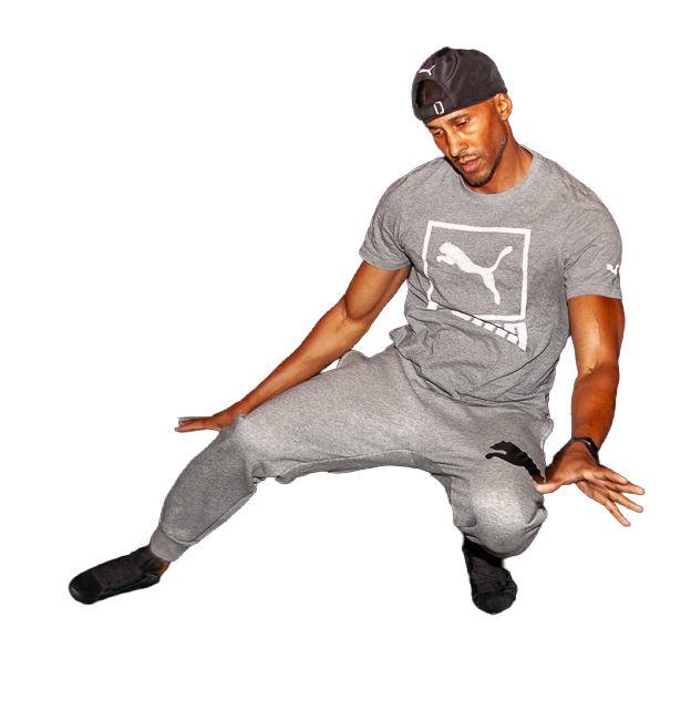 A man wearing a gray Puma t-shirt, gray sweatpants, and a black backward cap is crouching and balancing with one hand on the ground and the other hand near his knee, against a black background.