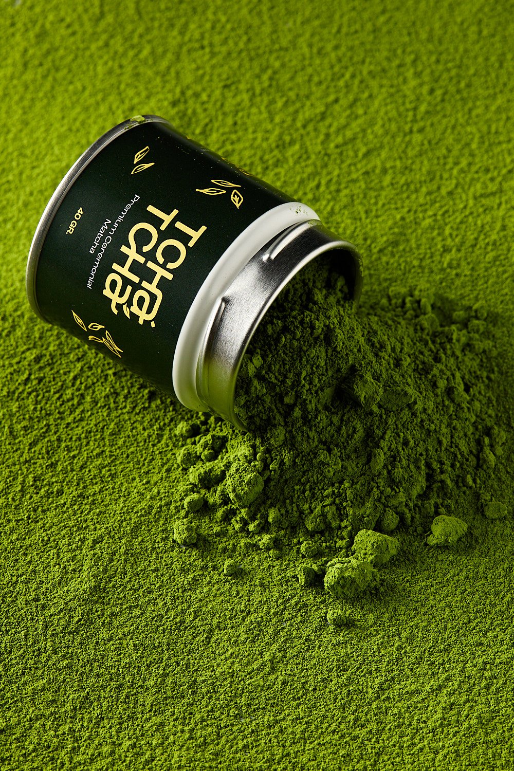 A black container labeled 'Matcha Tea' spills vibrant green matcha powder onto a matching green surface.
