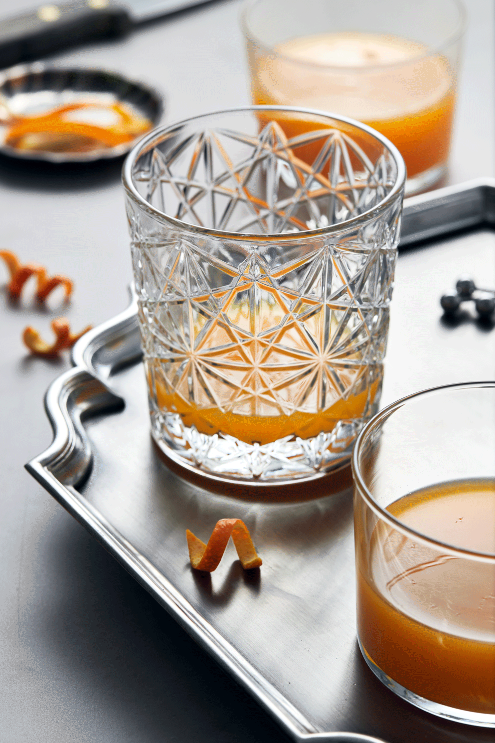 Close-up of a crystal glass with a patterned design, filled with orange-colored juice, placed on a silver tray with orange peel garnishes and additional glasses of juice