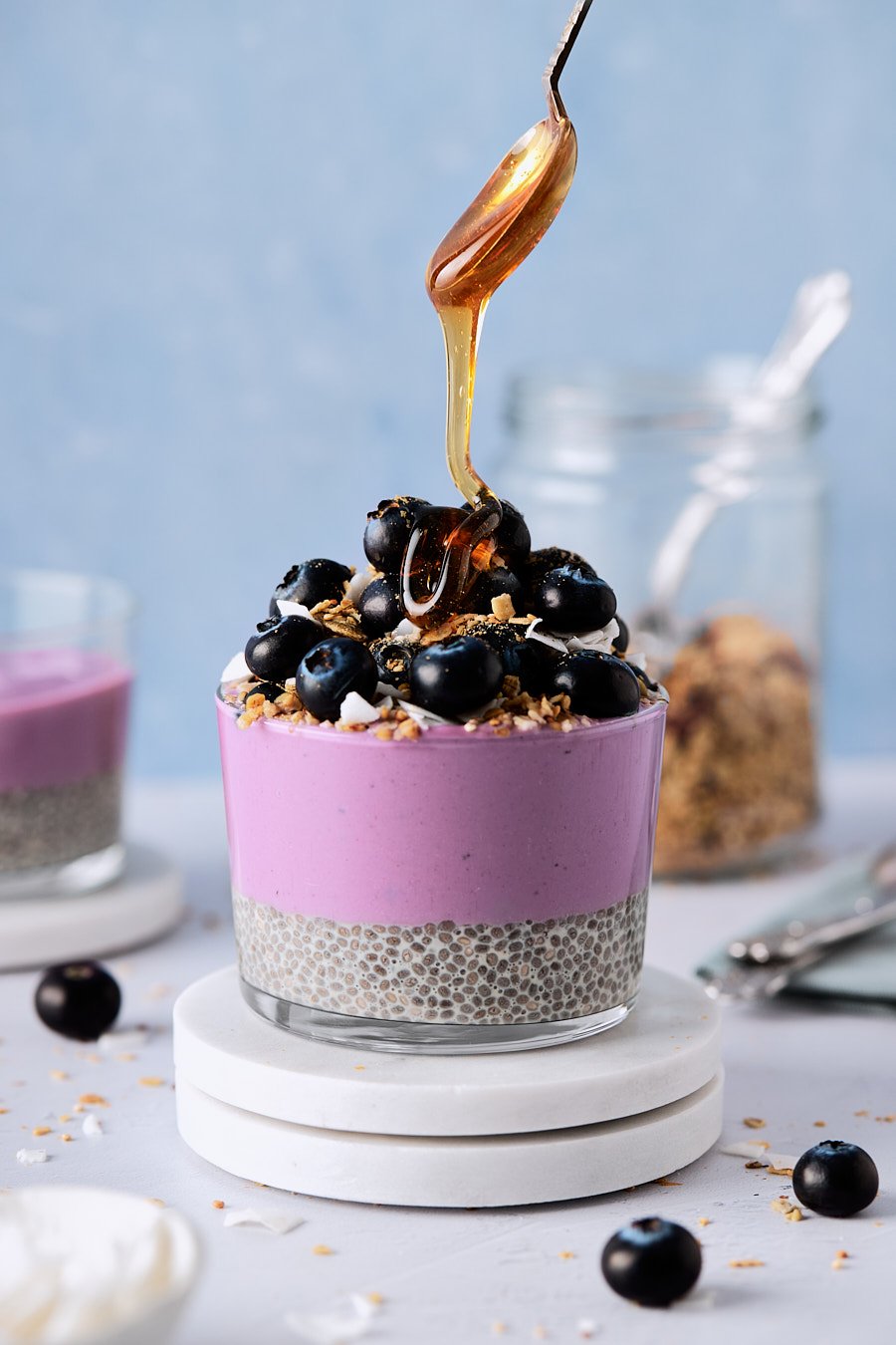 A layered dessert in a clear glass with a purple top layer, topped with blueberries, nuts, and syrup, on a white marble coaster.