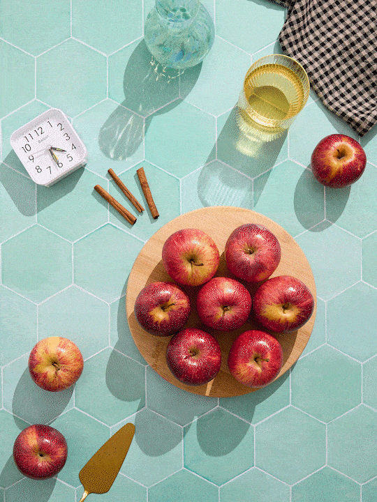 Top-down view of a kitchen countertop with a hexagonal tile pattern. There is a round wooden plate with eight red apples, a single apple outside the plate, a glass of yellow liquid, a small glass pitcher, a checkered cloth, a small alarm clock showin