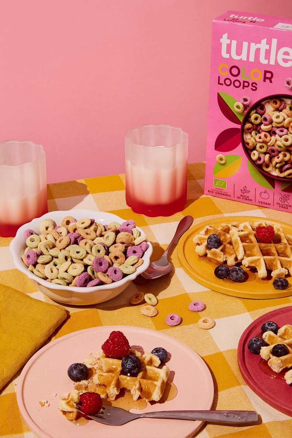Colorful breakfast scene with bowls of fruit and waffles, a box of multicolored cereal, glasses of pink-ombre juice, and plates with waffles topped with berries on a yellow checkered tablecloth.