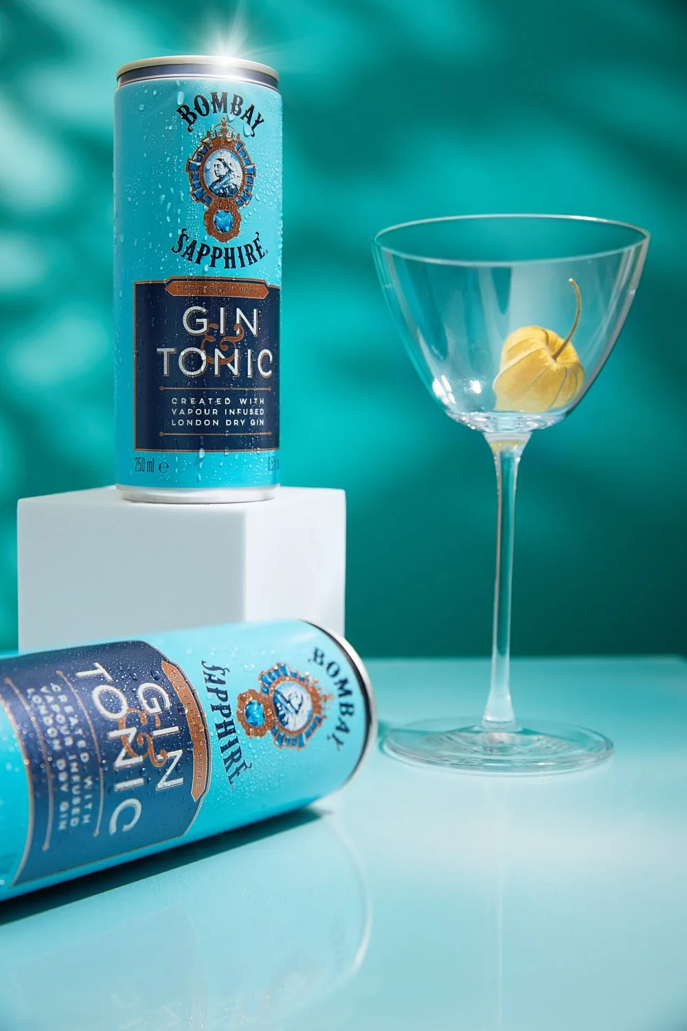 Two cans of Bombay Sapphire Gin Tonic with a lemon in a martini glass against a teal background. Drink Photography by Claudia Carmo. Lisbon, Portugal
