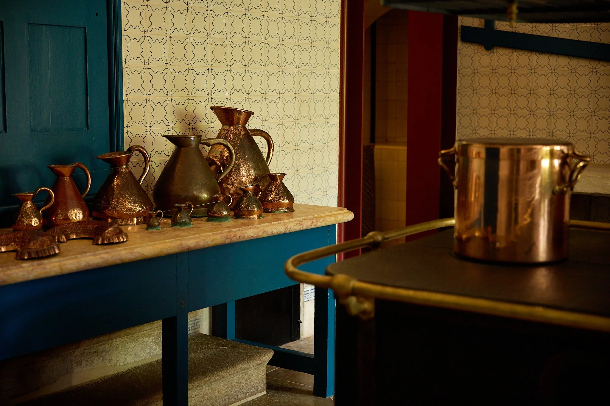Collection of copper pitchers displayed on a blue counter beside a large copper pot in a tiled interior