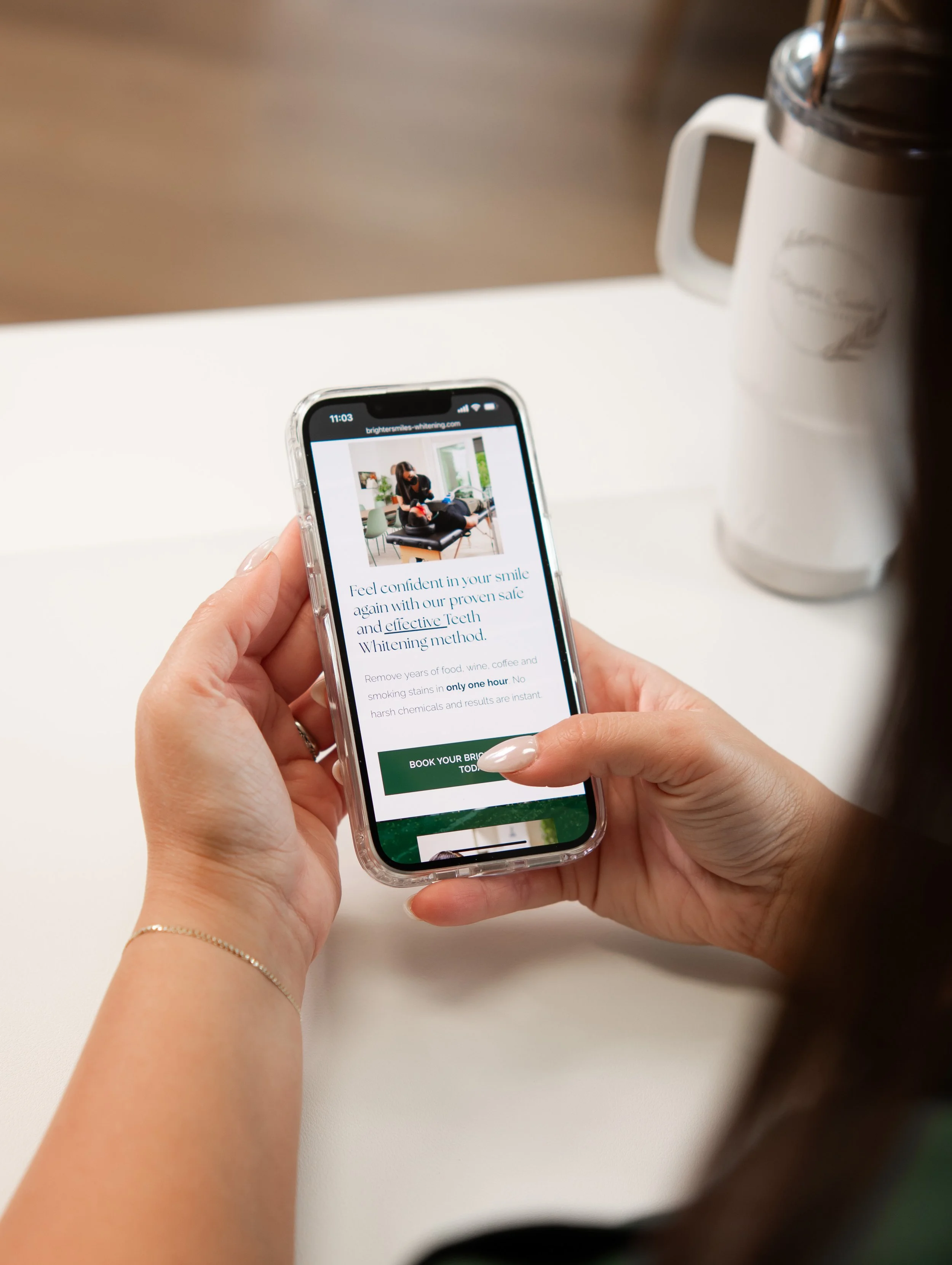 Person holding a smartphone displaying a teeth whitening service website, with a cup or pitcher on the table nearby.