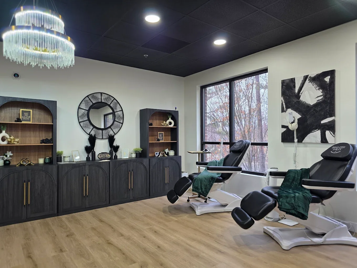 A skincare treatment room with two black reclining chairs, each with a green towel, in front of a large window. The room has light wood flooring, white walls, a black ceiling with recessed lighting, a decorative wall mirror, and black and wooden shelves with decor.