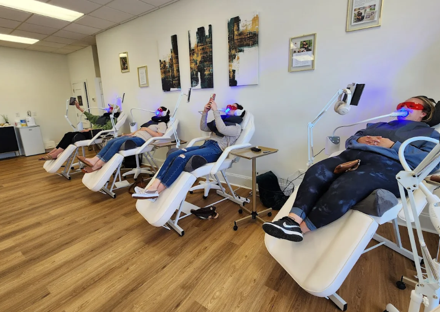 Four women at a skincare clinic lying in chairs with red LED masks on their faces and phones in their hands, receiving facial treatments.