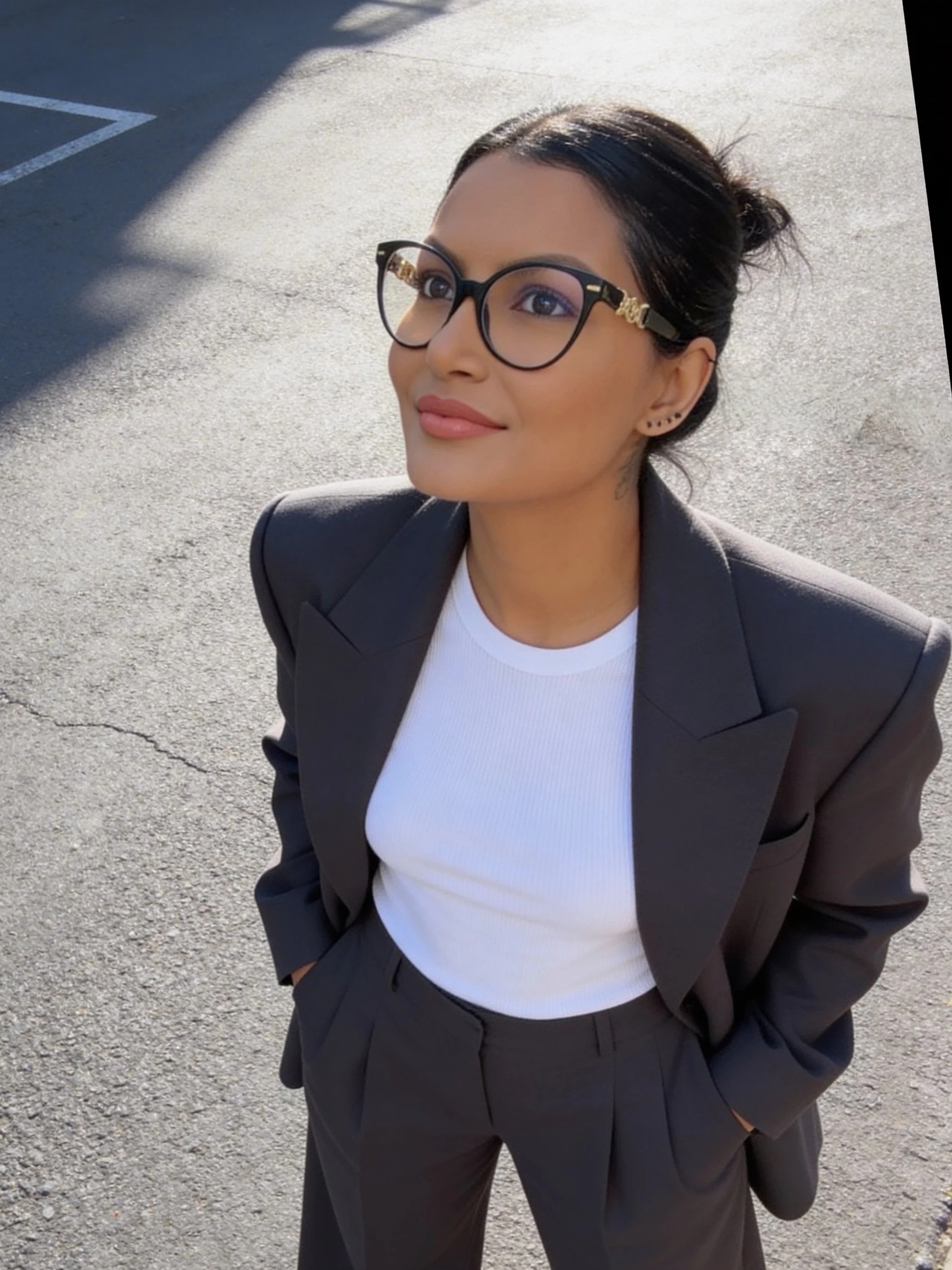 A young woman with glasses and earrings, wearing a white shirt and a black blazer, standing outdoors on pavement with her hands in her pockets, smiling gently.