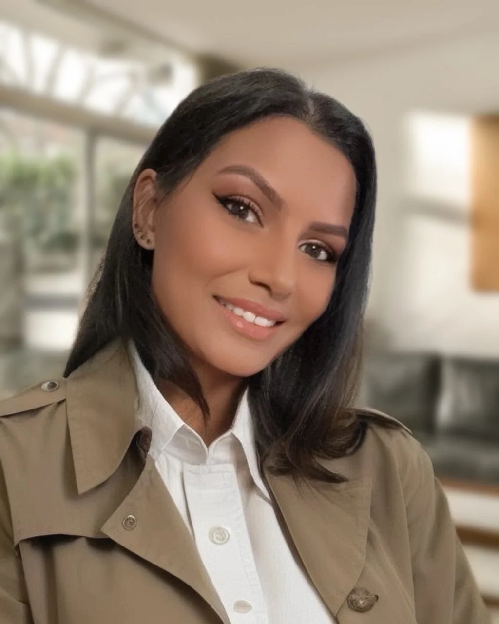 Woman with tanned skin smiling, she is wearing an olive coact with a white shirt underneath.