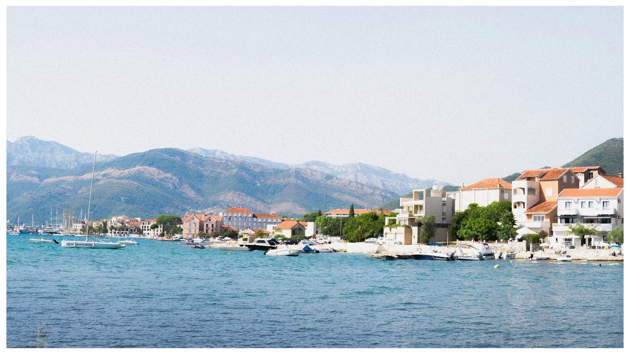 A vintage-style photograph of a coastal area of Tivat, Montenegro. You can see the sea, terracotta roofed houses and mountains in the distance.