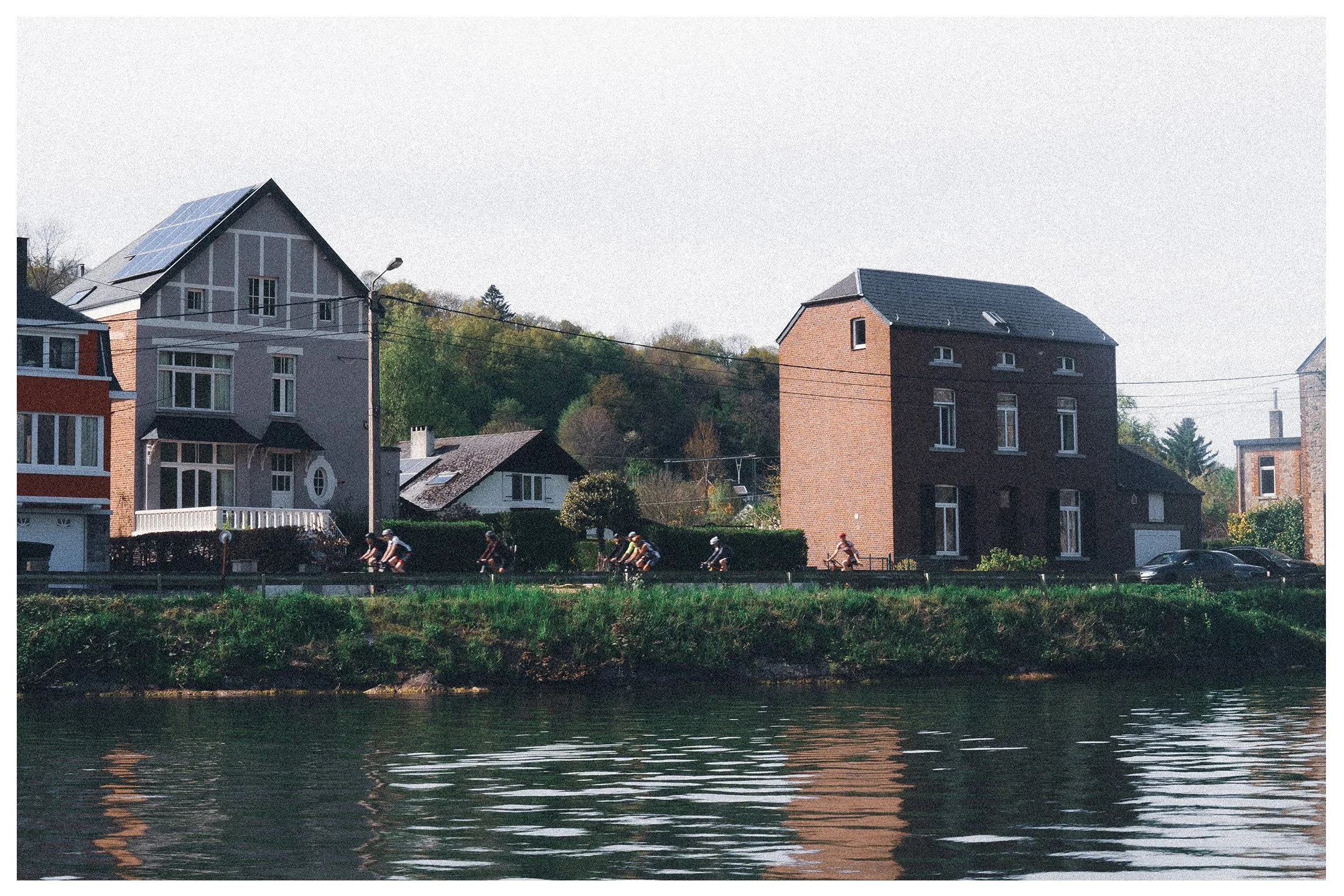 Colour photo taken on the river through Dinant, Belgium. The photo is looking at the shore, where 7 cyclists ride in single file next to traditional houses.