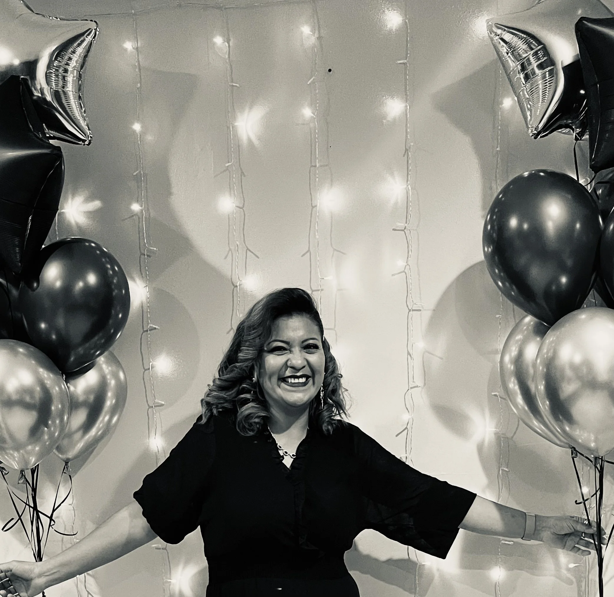 A woman with wavy hair smiling and posing with arms extended, surrounded by black and metallic balloons on a decorated wall with string lights.