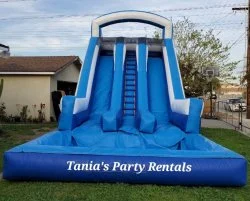 Large blue inflatable water slide with three lanes, labeled 'Tania's Party Rentals', outdoors in a yard with a house and trees in the background.