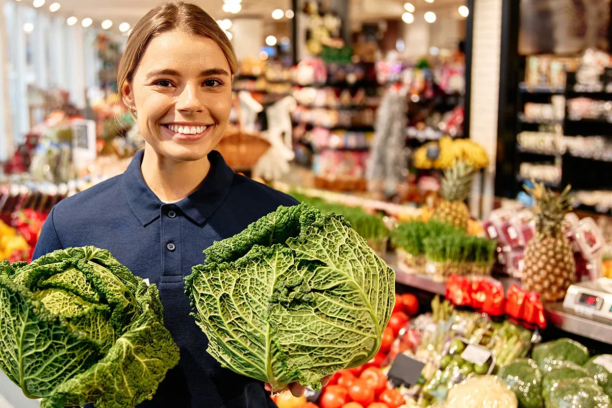 Smiling produce specialist holding fresh cabbages in a supermarket as part of a fresh produce product testing programme focused on quality, consistency, and consumer satisfaction.