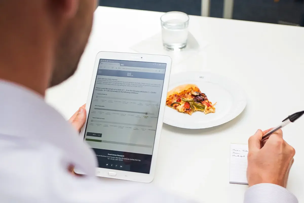 Person completing feedback on a tablet with a slice of pizza sample during product testing and benchmarking, showing how product testing and benchmarking evaluates taste, quality, and consumer preferences.