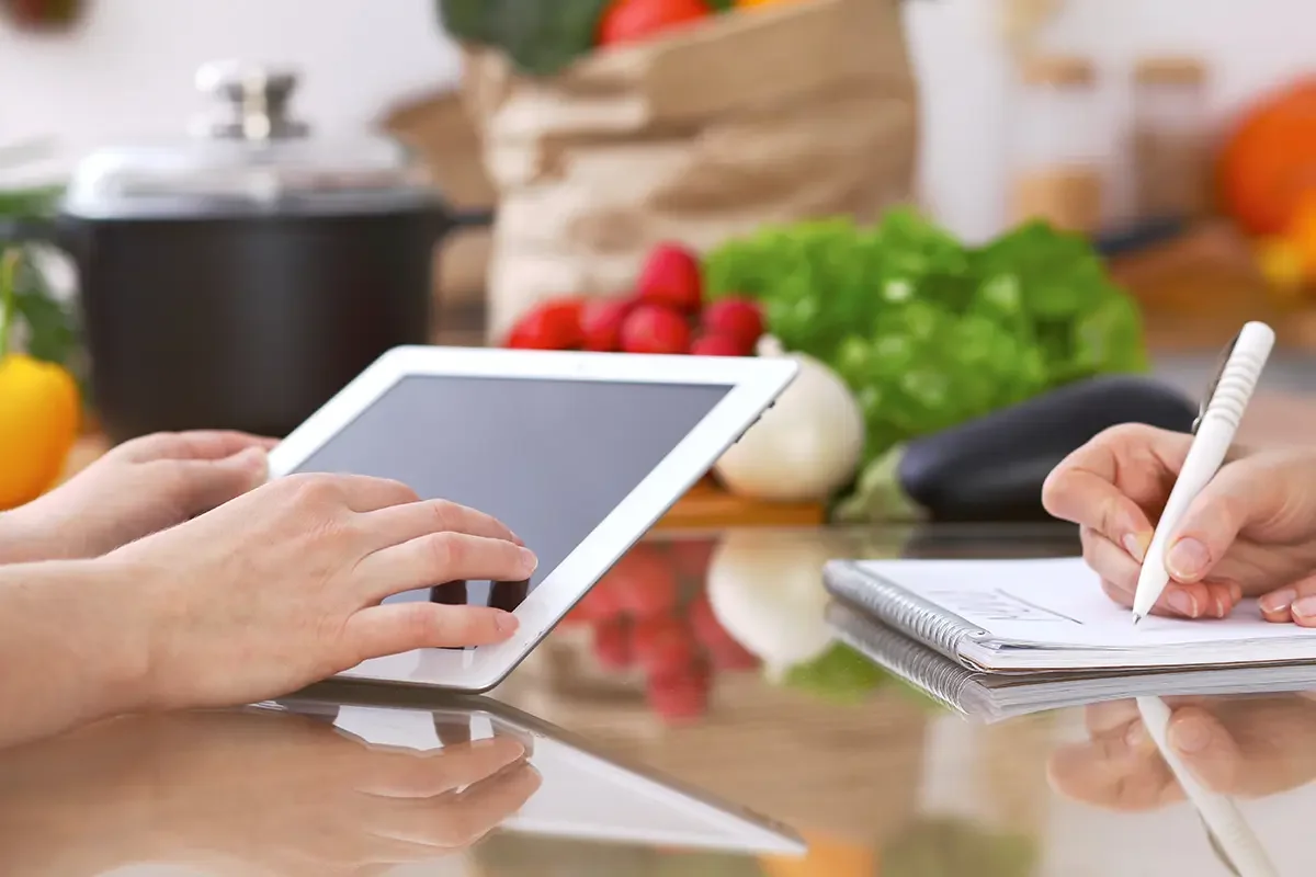 Hands using a tablet and taking notes in a kitchen setting with fresh produce, symbolising research, analysis, and decision-making in product testing and benchmarking