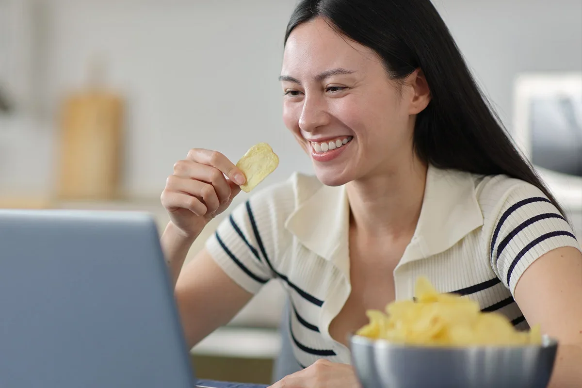 Smiling woman taking part in an online session while eating crisps, representing how Consumer Closeness Research UK connects brands with real consumers in natural moments to generate authentic insights and deeper understanding.