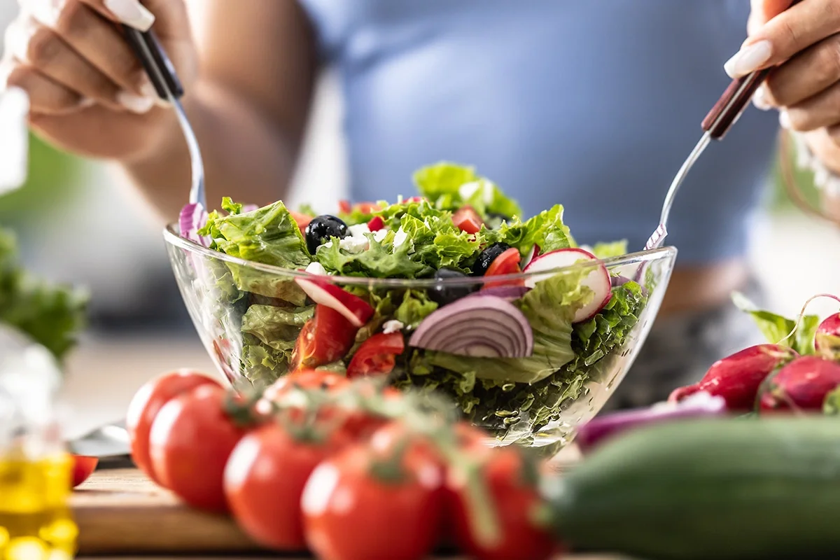 Person preparing a vibrant salad with tomatoes, lettuce, onions, and olives, representing the importance of a fresh produce consumer testing programme in understanding shopper preferences, improving product quality, and enhancing taste appeal.