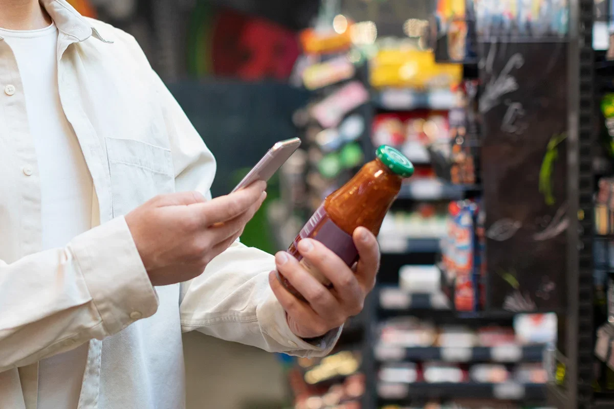Shopper scanning a sauce bottle with a smartphone in a supermarket, representing how Customer journey research UK tracks digital and in-store behaviours to understand decision-making, touchpoints, and purchase influences.