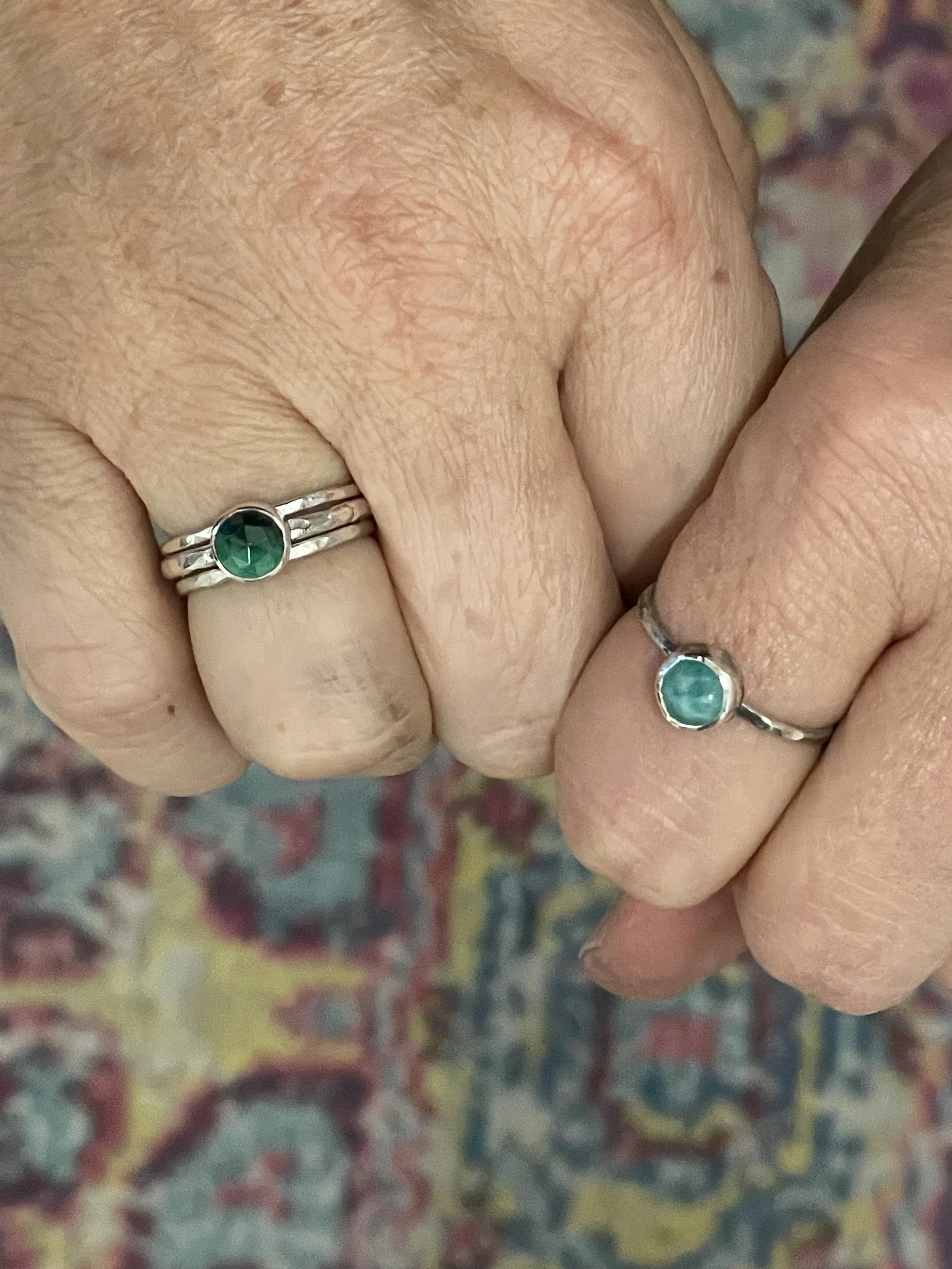 Two hands adorned with elegant stacking rings, each featuring a single bezel-set stone. These handmade rings highlight the beauty of minimalist design and fine craftsmanship