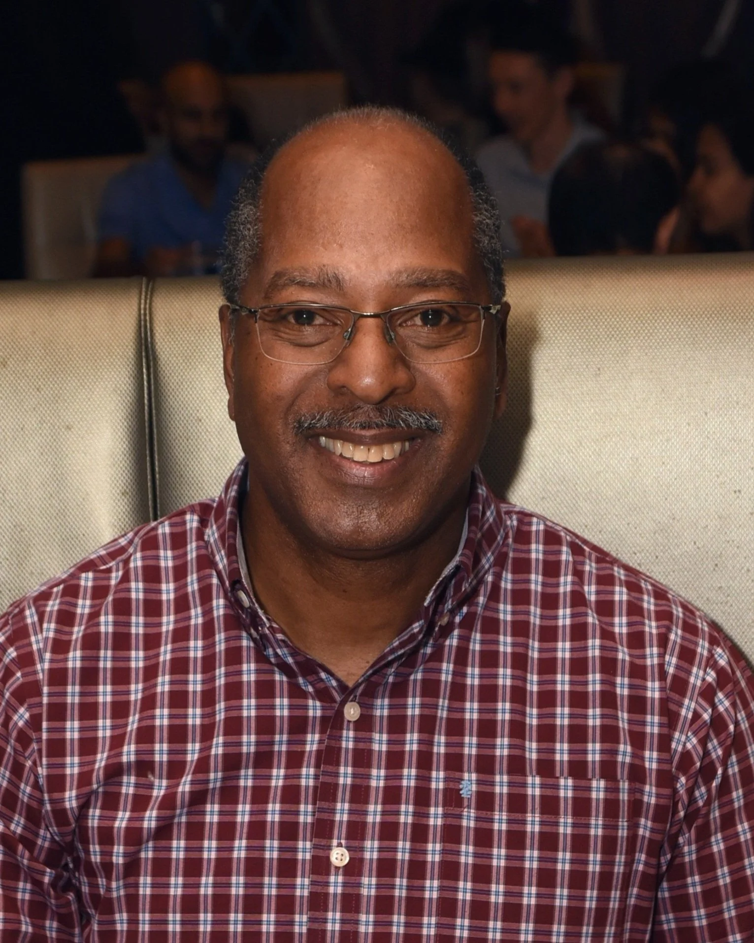 Smiling man wearing glasses and a red plaid shirt, seated indoors.